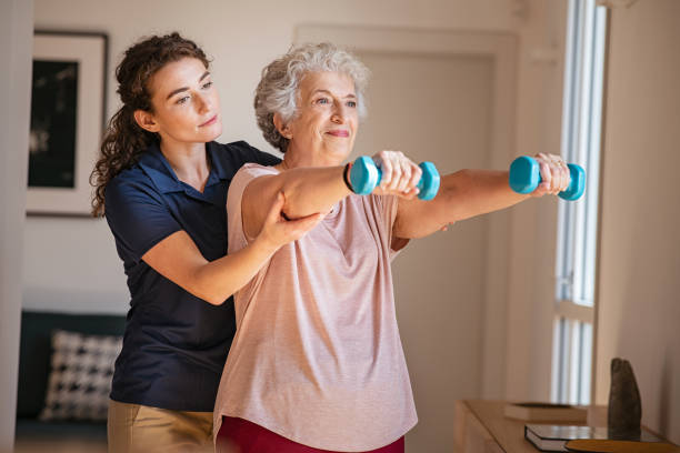 Old woman training with physiotherapist using dumbbells at home. Therapist assisting senior woman with exercises in nursing home. Elderly patient using dumbbells with outstreched arms in a physical therapy session in clinic.